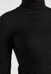 Nahaufnahme einer Person, die einen schwarzen, langärmligen Pullover mit Rollkragen trägt, wobei die Textur und Passform über Schulter und Brust deutlich zu sehen sind.