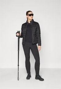 Giacca nera isolata con motivo trapuntato, abbinata a un top nero a maniche lunghe e pantaloni. Tiene bastoni da trekking e indossa calzature nere.