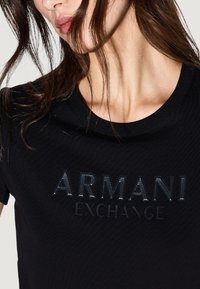Mujer con una camiseta negra de Armani Exchange con un logo texturizado, el cabello parcialmente cubriendo la cara, visible desde el cuello hasta la parte superior del pecho.