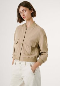 Light beige bomber jacket with ribbed cuffs and hem, featuring two large front pockets and a snap button closure, made of a smooth fabric.