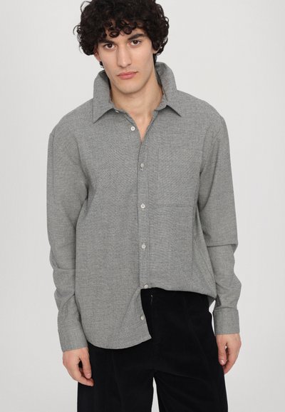 Cămașă gri, cu nasturi, dintr-un material texturat, cu guler răspândit, un singur buzunar pe piept și mâneci lungi; purtată cu pantaloni din corduroy închis la culoare.