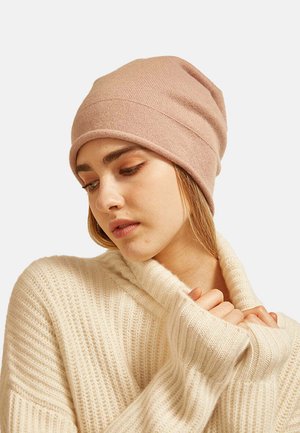 Un berretto lavorato a maglia di colore rosa chiaro presenta un materiale morbido e elastico con una leggera costina. Il modello indossa un maglione crema a trecce, con le mani giunte.