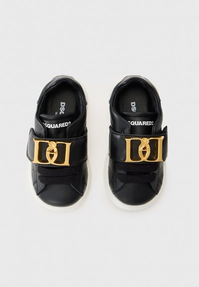 Svarta lädersneakers med en vit sula. Har ett brett band med en design av ett guldspänne och svarta snören. Varumärkeslogotypen synlig inuti.