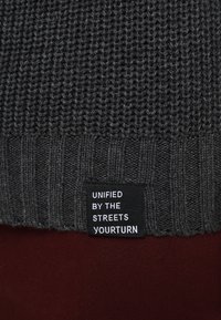 Dunkelgrauer Strickstoff mit einem schwarzen Etikett, auf dem in weißer Schrift "UNIFIED BY THE STREETS YOURTURN" steht.