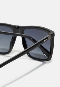 Black sunglasses with angular frames, dark gradient lenses, and metallic accents. Features brand logo on the side. Textured finish on temples.