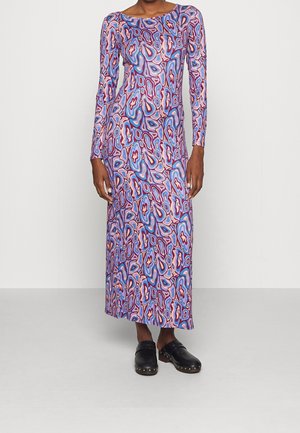 Femme portant une robe longue à manches longues, jusqu'aux chevilles, avec un motif abstrait multicolore et des sabots noirs à enfiler ornés de clous.