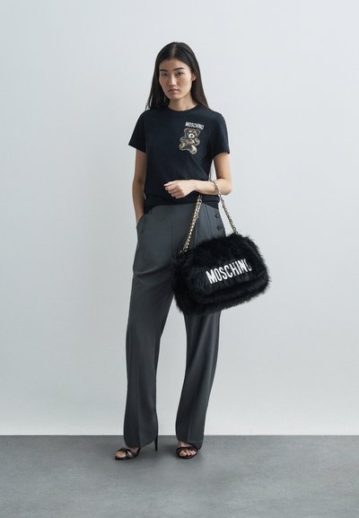 Mulher vestindo uma t-shirt preta da Moschino com um desenho de urso, calças cinzas, saltos pretos, segurando uma mala de mão peluda preta da Moschino com alça de corrente.