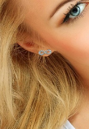 Boucles d'oreilles en argent en forme d'infini avec des strass transparents, présentant une finition polie et des détails complexes, portées avec des cheveux blonds.
