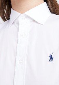 White button-up shirt featuring a pointed collar, clear plastic buttons, and a dark blue embroidered logo on the left chest. Smooth texture.