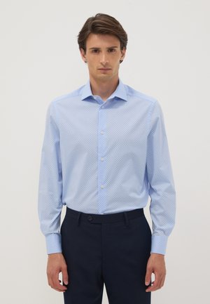 Mann trägt ein hellblau gemustertes langärmeliges Hemd, das in dunkelblaue Hose gesteckt ist, und steht vor einem schlichten hellen Hintergrund.