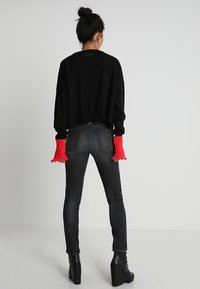 Schwarzer, cropped Pullover mit geripptem Saum, kombiniert mit figurbetonten, dunkelgrauen Jeans. Rote, fransige Ärmelakzente und schwarze Plateaustiefel sind sichtbar.