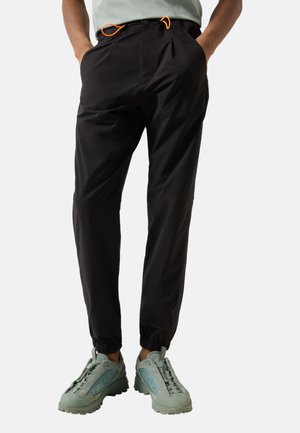 Osoba nosząca czarne spodnie z elastycznymi mankietami i pomarańczowymi sznurkami, ręce w kieszeniach oraz zielone sneakersy na białym tle.