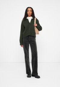 Cardigan en tricot vert foncé avec un col et une fermeture à boutons, associé à un pantalon en velours côtelé noir et des chaussures à plateforme noires. Sac fourre-tout beige.