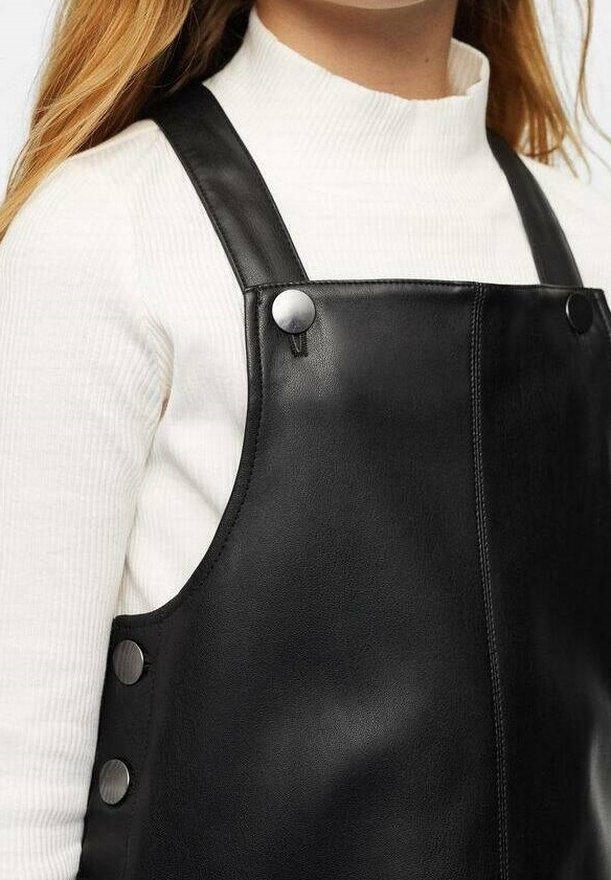 Femme portant un pull à col roulé côtelé blanc sous une robe salopette en simili cuir noir avec des boutons-pression argentés sur les bretelles et les côtés.