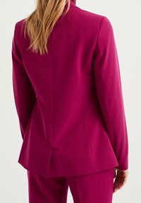 Tajmad magenta kavaj med slagkrage, långa ärmar, slätt tyg och en sprund bak, kombinerad med matchande byxor.