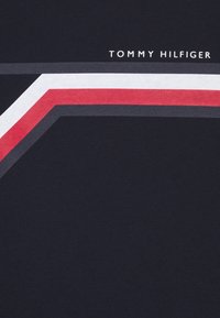 Navy t-shirt featuring a horizontal stripe design in red, white, and gray. "TOMMY HILFIGER" logo printed in white above the design.