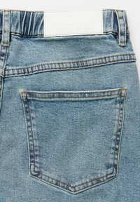 Light blue denim jeans featuring a back pocket with orange stitching and a white leather label at the waistband with embossed lettering.