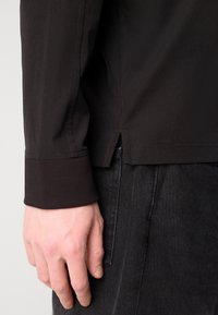 Fekete, hosszú ujjú póló bordázott mandzsettával és oldalsó hasítékkal, sima anyagból készült. Kéz sötét, texturált anyagon pihen.