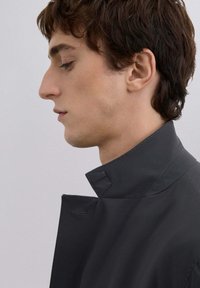 Abrigo negro de cuello alto con una textura suave, que presenta un diseño minimalista y un ajuste a medida. Las costuras visibles acentúan el detalle del cuello.