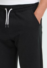 Nahaufnahme einer Person, die schwarze Jogginghosen mit weißen Kordeln trägt, wobei die Hand in die rechte Tasche steckt.