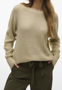 Beżowy sweter dzianinowy o fakturowanym wzorze, z żebrowanym dekoltem i lekko luźnym krojem, zestawiony z oliwkowymi spodniami z ściągaczem.
