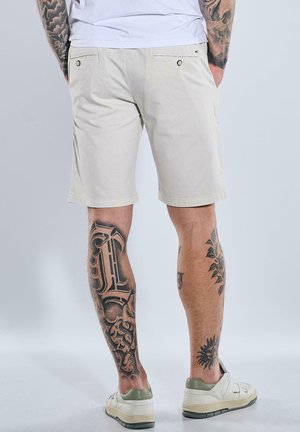 Helle beige Shorts aus Baumwolle mit glatter Textur, ausgestattet mit zwei hinteren Taschen und dezenten Nähdetails. Getragen mit weißen Sneakers.