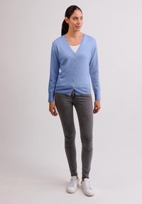 Ljusblå stickad cardigan med V- ringning, knappfront, långa ärmar och ribbad nederdel, kombinerad med grå byxor och vita sneakers.