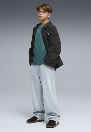Chłopiec stojący z rękami w kieszeniach, ubrany w oversize'ową czarną kurtkę, turkusową koszulę, obszerne jasne jeansy i czarne trampki na szarym tle.