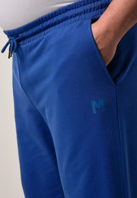 Blå sweatpants gjorda av mjukt tyg, med elastisk midjeband, sidofickor och en diskret logotyp i en mörkare nyans av blått.