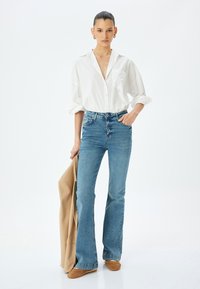 Camisa blanca de botones con mangas enrolladas, jeans acampanados azules y americana color beige en la mano. Unos zapatos marrones completan el conjunto. Diseño y corte simples.