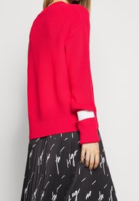 Pull en tricot rose vif avec une texture côtelée et un accent blanc au niveau du poignet, associé à une jupe plissée noire arborant un motif textuel blanc.
