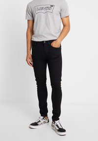 Jeans ajustados negros combinados con una camiseta gris de Levi's con un logo negro, complementados con zapatillas de cuadros en blanco y negro.