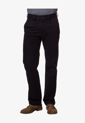 Man die zwarte rechte broek draagt, een grijs overhemd in de broek gestopt en bruine veterschoenen, staand tegen een witte achtergrond.