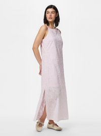 Femme portant une robe maxi sans manches rose clair avec des détails en œillets floraux et une fente latérale, associée à des chaussures plates beige, debout de côté.