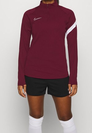 Vrouw die een bordeauxkleurige lange mouwen sporttop met witte swoosh draagt, zwarte korte broek en witte kniehoge sportsokken, staand tegen een effen achtergrond.