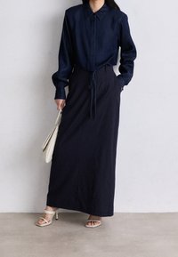Camicia blu navy con colletto, abbinata a una lunga gonna scura con vita a striscia. La modella tiene una borsa leggera e indossa tacchi a spillo.