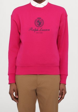 Osoba nosząca jasno różową bluzę Ralph Lauren na białej koszuli z kołnierzykiem i beżowych spodniach, stoi na białym tle.