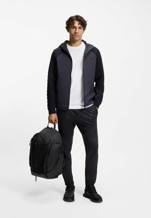Dunkelgraue Jacke mit strukturierten Ärmeln, weißes T-Shirt, schwarze Hose mit dezentem Muster und ein schwarzer Rucksack mit Reißverschlüssen, in der Hand gehalten.