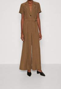 Person som bär en brun bältevid baddräkt med korta ärmar och svarta spetsiga blockklackar, stående mot en enfärgad bakgrund.