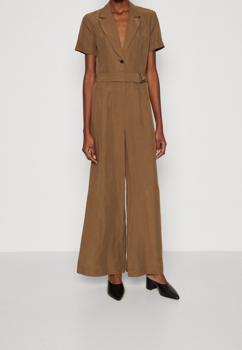 Person som bär en brun bältevid baddräkt med korta ärmar och svarta spetsiga blockklackar, stående mot en enfärgad bakgrund.