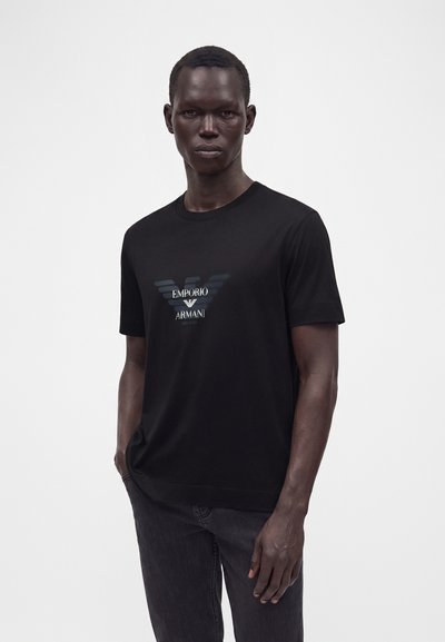 Άνδρας που φοράει μαύρο μπλουζάκι Emporio Armani με εκτύπωση λογοτύπου, στέκεται με το ένα χέρι στην τσέπη μπροστά σε λευκό, μονόχρωμο φόντο.