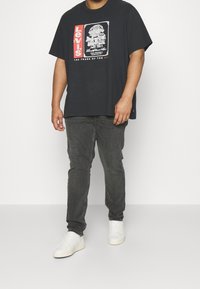 Homme portant un t-shirt graphique Levi's noir, un jean gris foncé et des baskets blanches, debout devant un fond blanc uni.