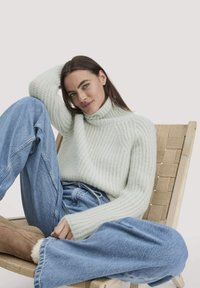 Frau, die auf einem geflochtenen Holzstuhl sitzt, ein weißes Strick-Turtleneck-Sweatshirt, lockere blaue Jeans und braune Lammfellstiefel trägt und mit dem Arm hinter dem Kopf posiert.