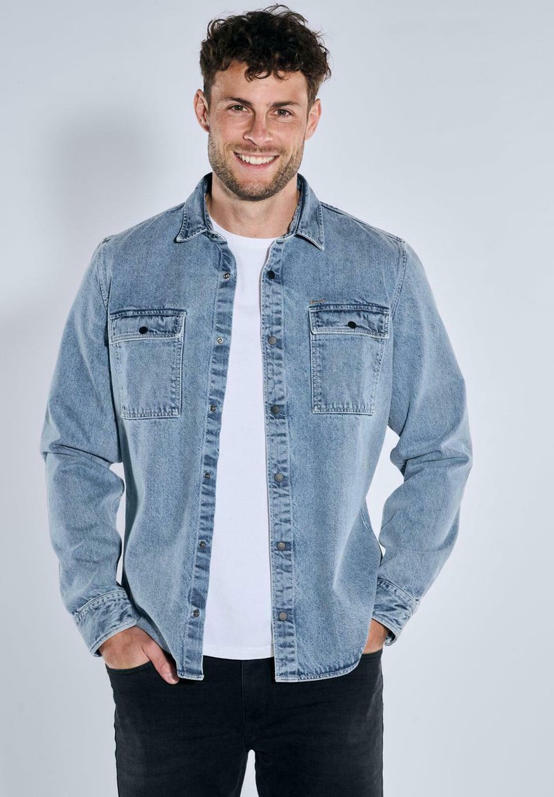 Hellblaues Denimhemd mit Knopfverschluss, zwei Fronttaschen und klassischem Kragen. Getragen über einem weißen T-Shirt, kombiniert mit schwarzen Hosen.