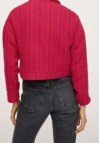 Przycięta, teksturowana kurtka w jaskrawym różu z delikatnym wzorem, zestawiona z czarnymi spodniami dżinsowymi z wysokim stanem i tylnymi kieszeniami.