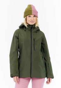 Vrouw die een half olijfgroen, half roze gebreid mutsje, een olijfgroene sweatshirt met rits en capuchon, en roze broeken draagt tegen een witte achtergrond.