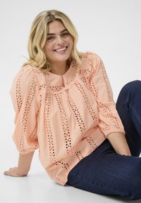 Peach-colored blouse with a textured design featuring lace cutouts, a rounded neckline, and three-quarter sleeves, styled with dark blue jeans.