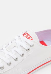 Zapatillas de lona blancas con ojales de metal, cordones blancos y un logo rojo en la lengüeta. Acentos rojos en la suela. Textura suave.