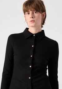 Zwarte langemouwen shirt met ribbelstructuur, voorzien van een klassieke kraag en gouden knopen aan de voorkant. Eenvoudig en op maat gesneden ontwerp.