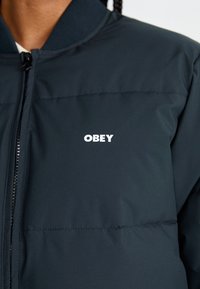 Giubbotto imbottito di un blu navy scuro con colletto a costine, zip frontale e logo bianco "OBEY" nella zona sinistra del petto. Texture liscia e isolata.